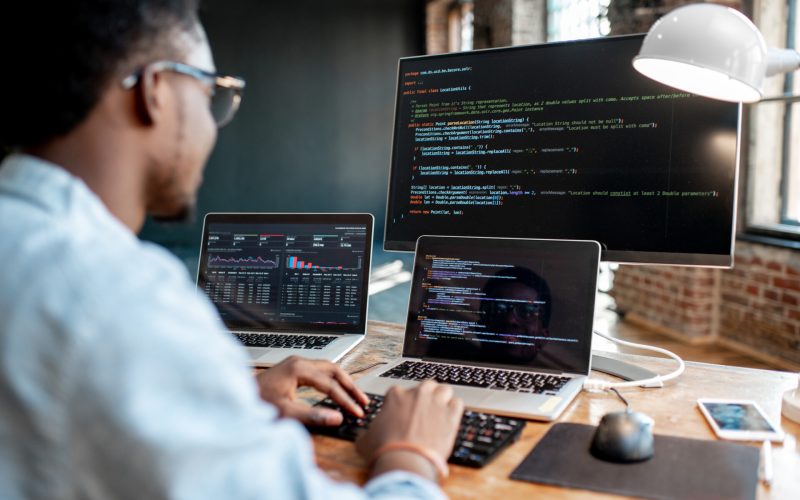 Young african male programmer writing program code sitting at the workplace with three monitors in the office. Image focused on the screen